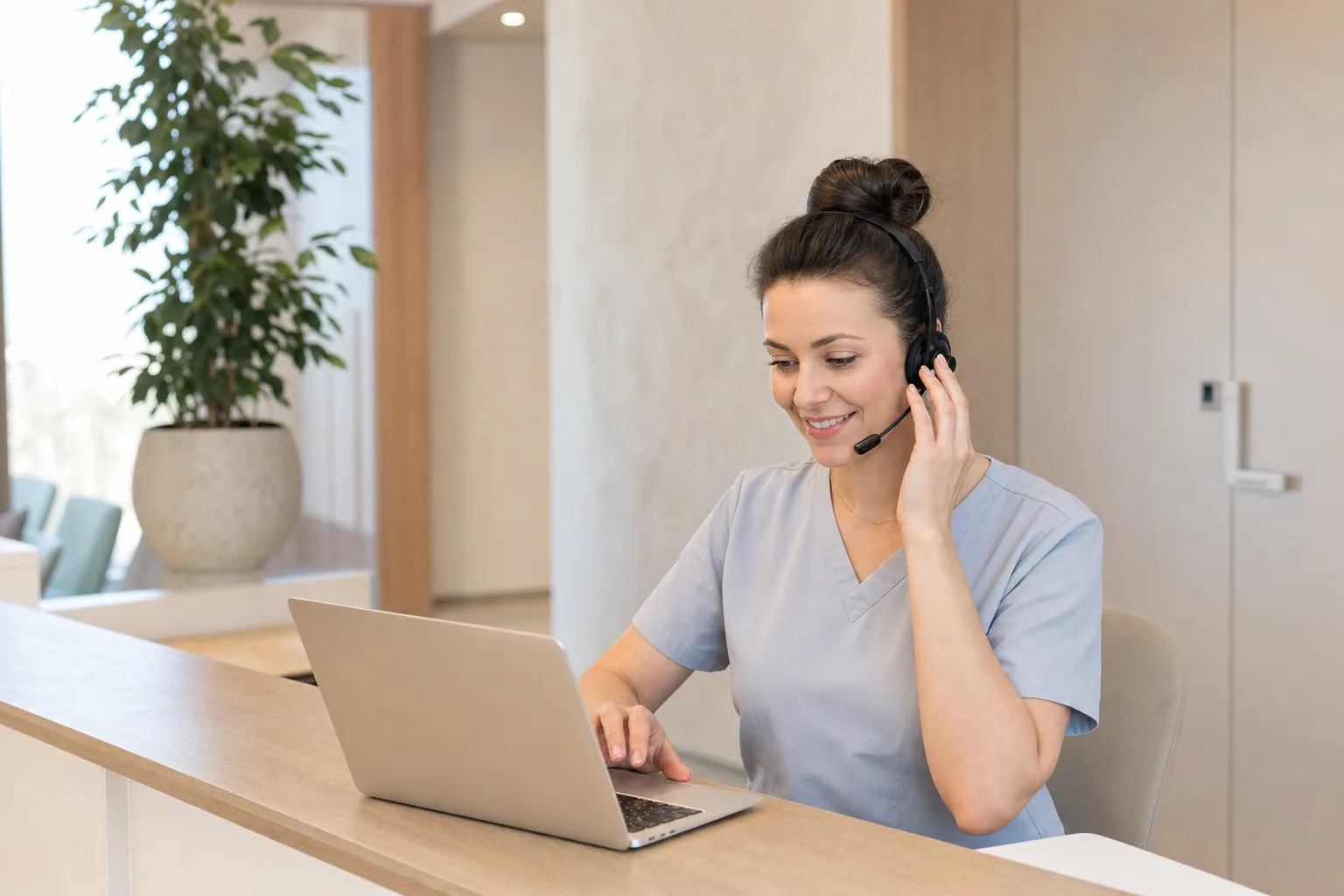 Receptionist in scrubs at a modern clinic front desk smiling on a phone headset.