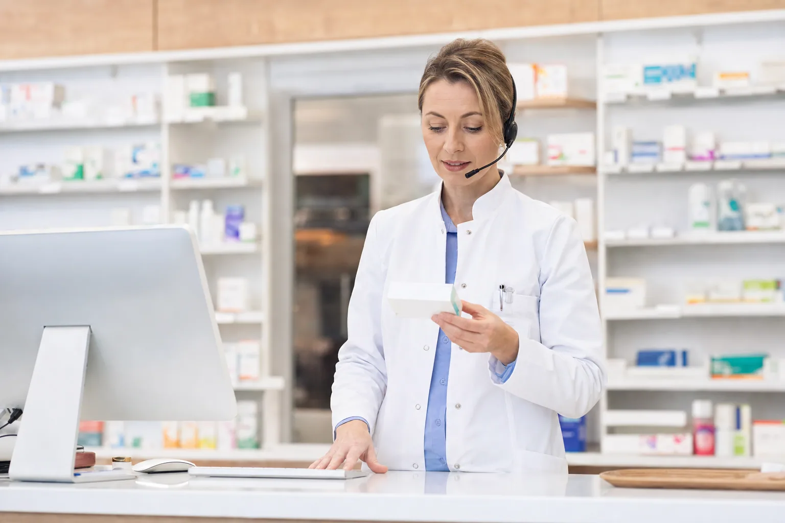 Pharmacist behind a clean counter holding a medication box and a phone headset.
